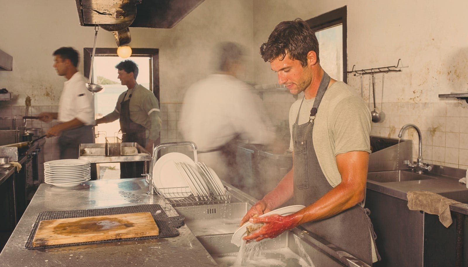 Young dishwasher working inside a busy 1970s-style seafood restaurant kitchen